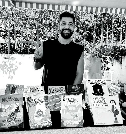 Foto em preto e branco de Felipe Manhães, vestido de camiseta sem manga, fazendo V com os dedos e usando pulseira de couro. Está de pé atrás de uma barraca com diversas revistas em quadrinhos à sua frente. Ao fundo, um muro com plantas e uma grade metálica.